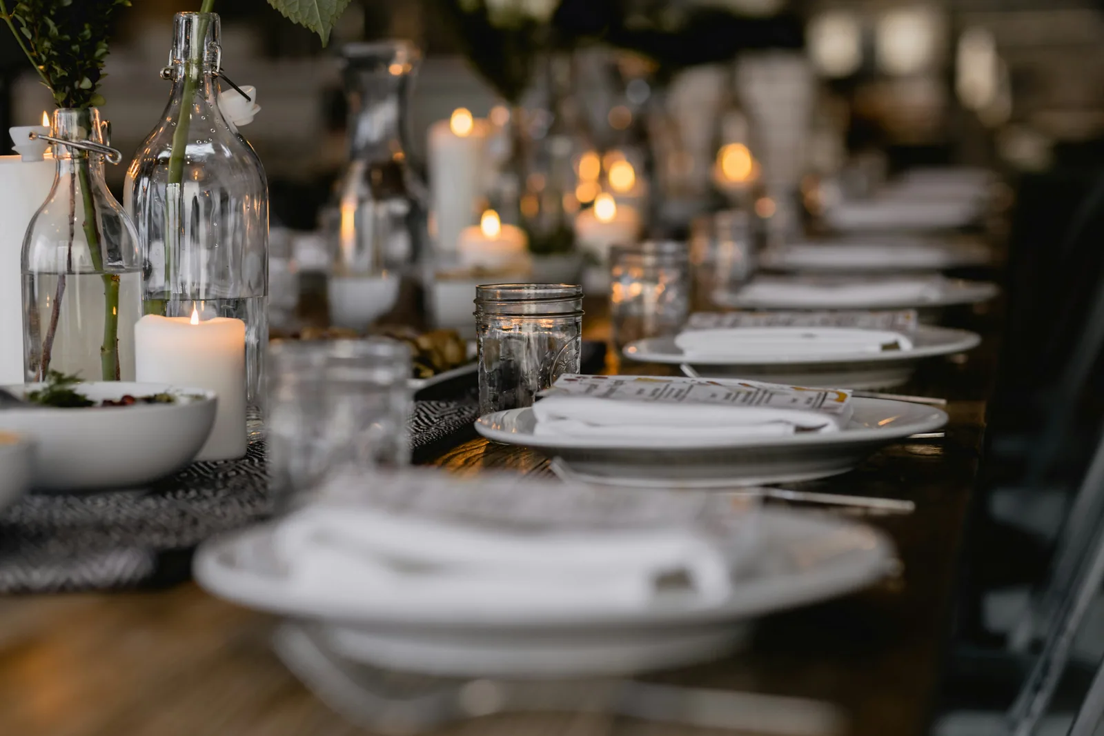A long dining table set with white plates, candles, and glass bottles