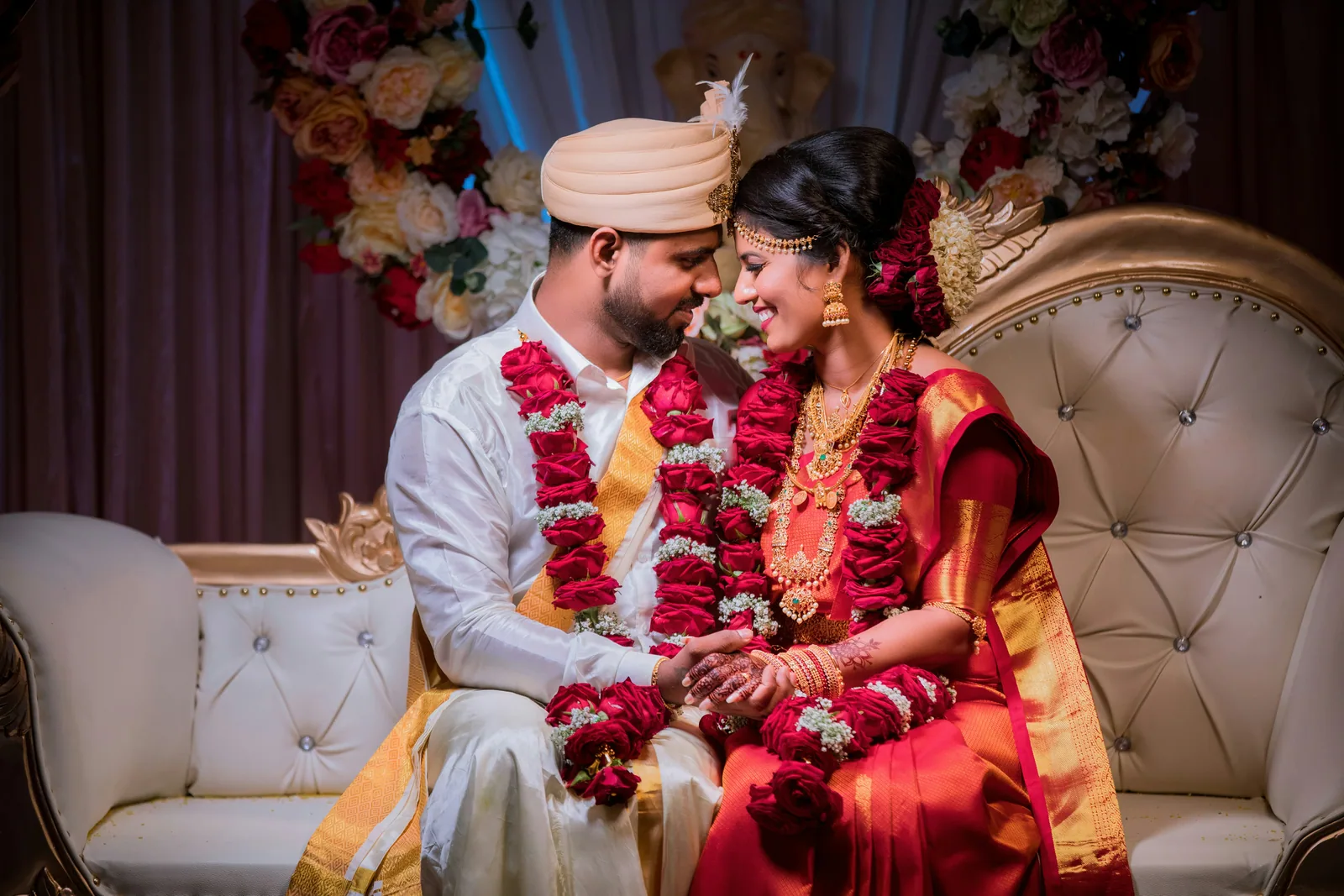 A couple in red and gold Indian wedding clothes, with garlands, on a carved sofa