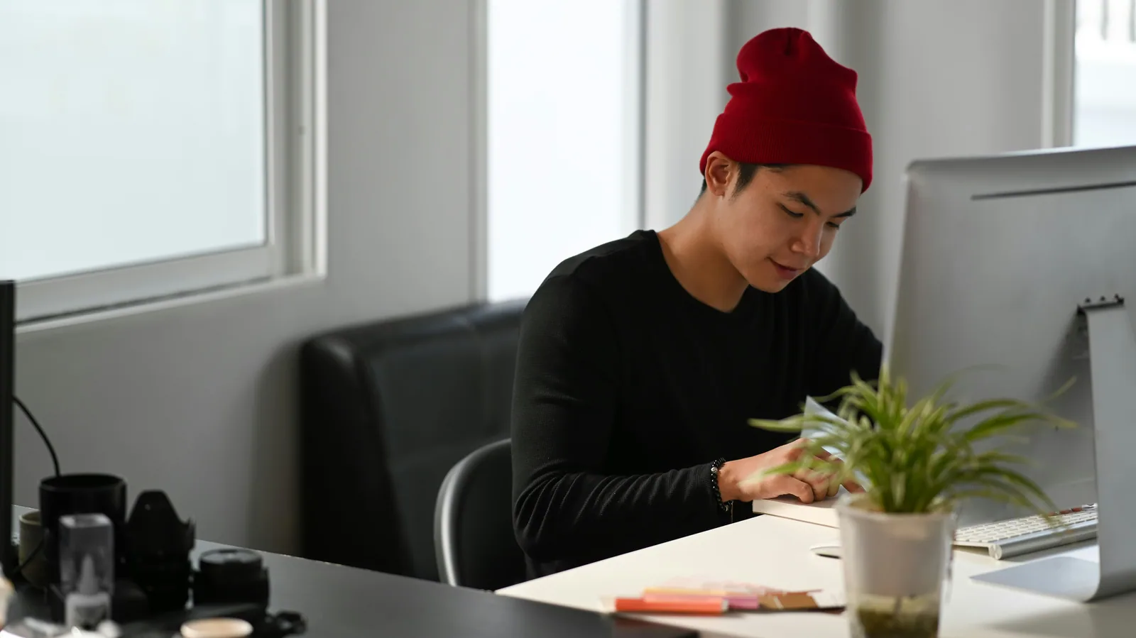 A designer in a red beanie working at a white desk with a plant