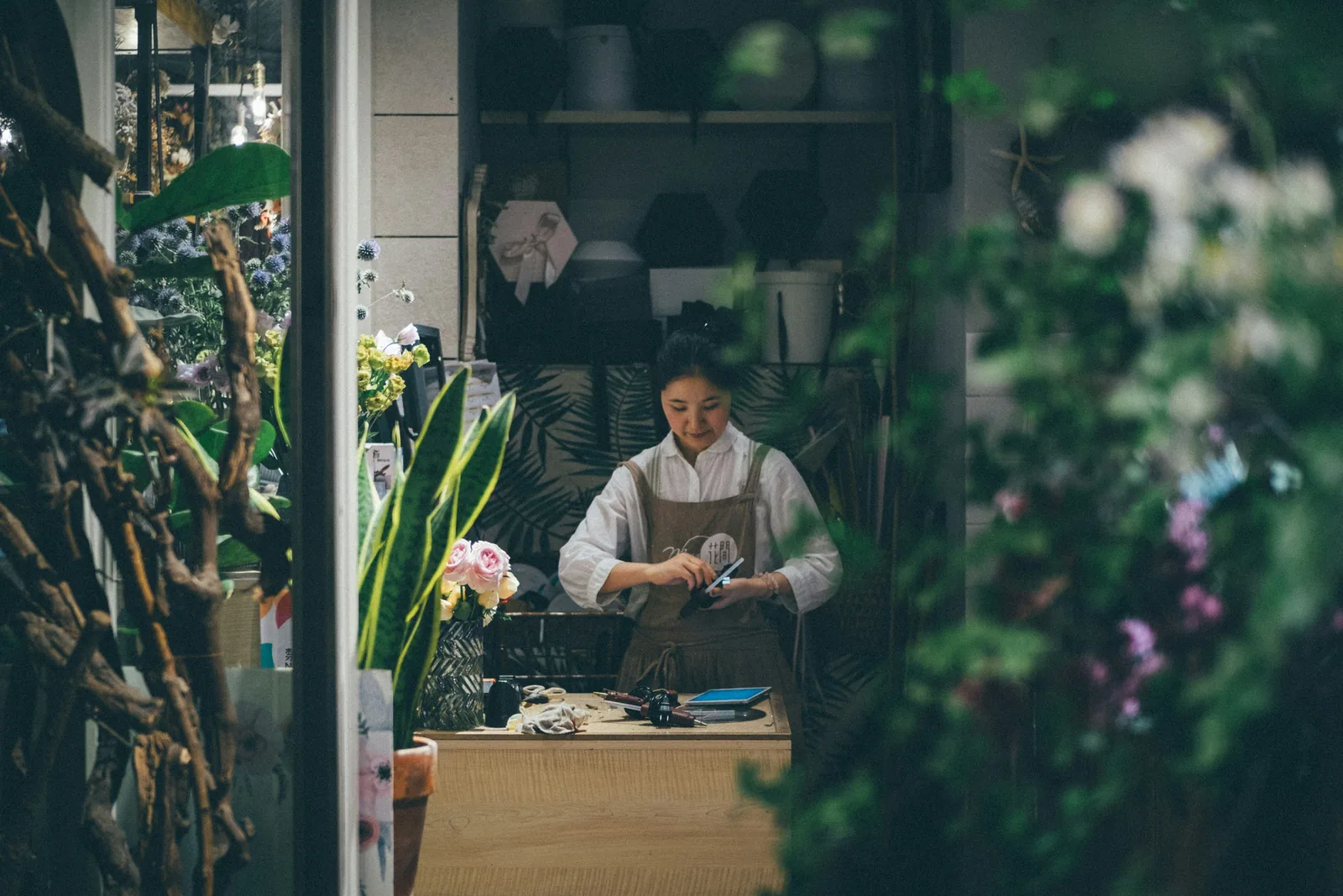 A florist at her workbench, seen through the plants of the shop