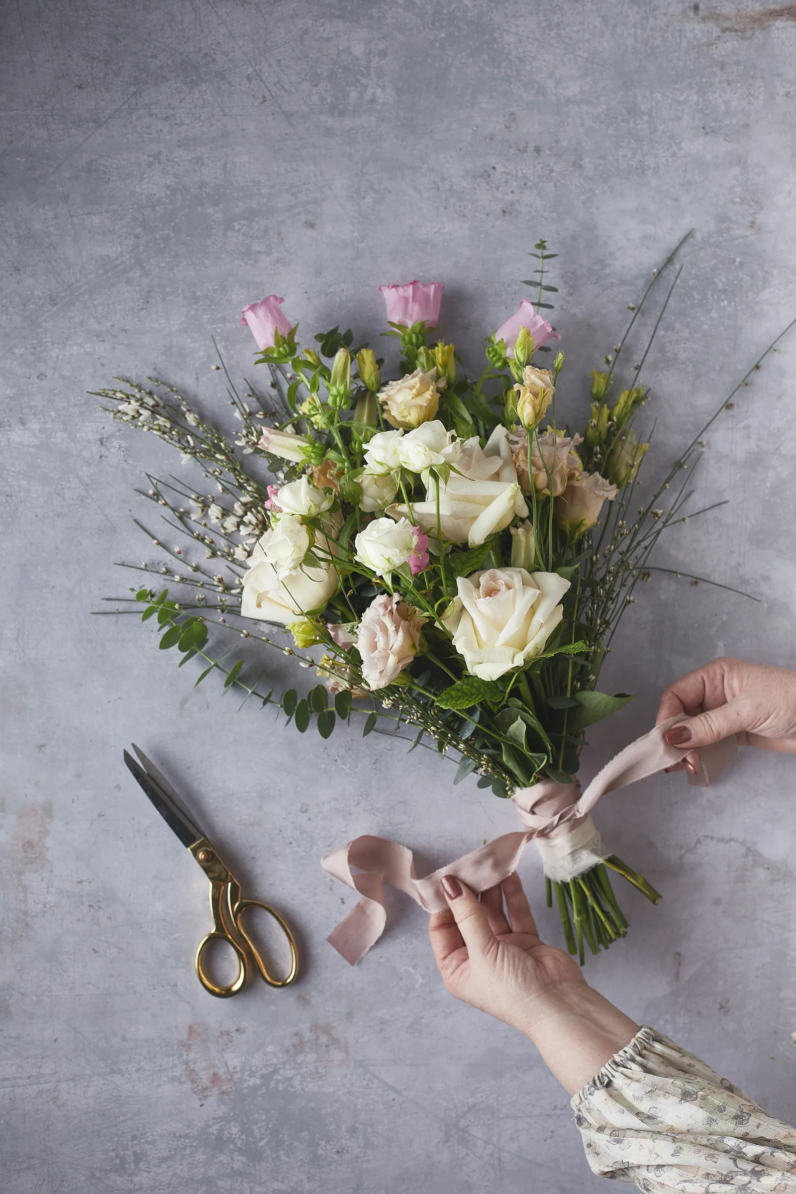 Hands tying a white bouquet with a pale ribbon, scissors beside it