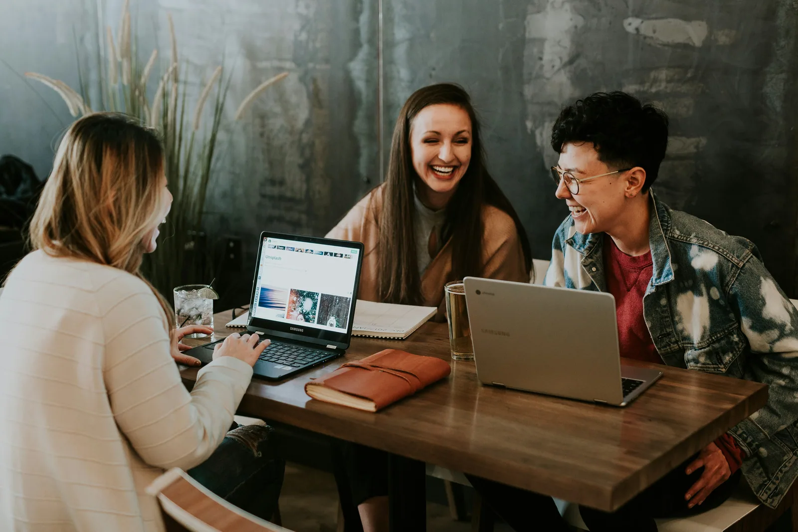 Three colleagues laughing together at a wooden table with laptops