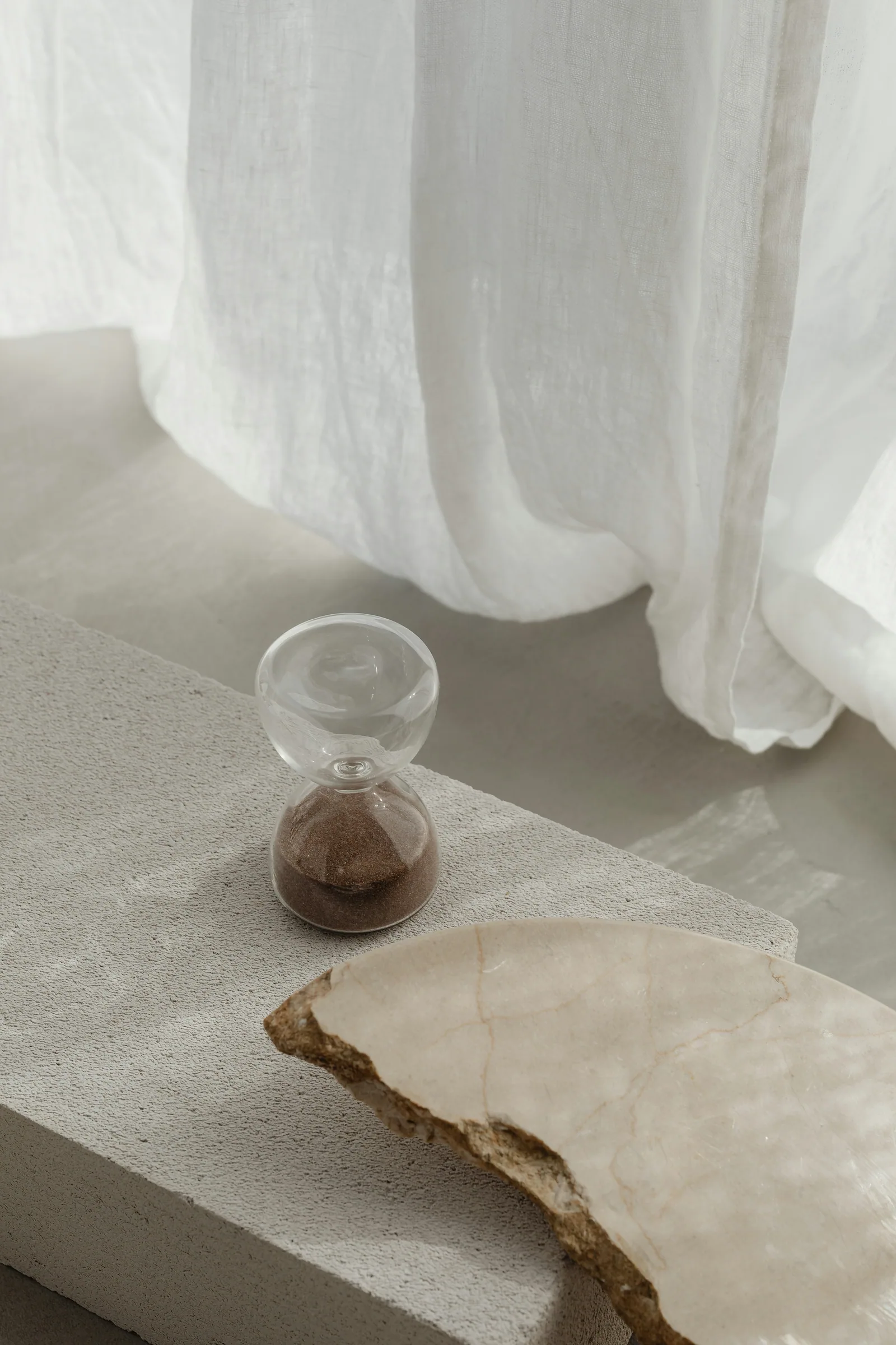 An hourglass and a piece of stone on a plaster ledge by sheer curtains