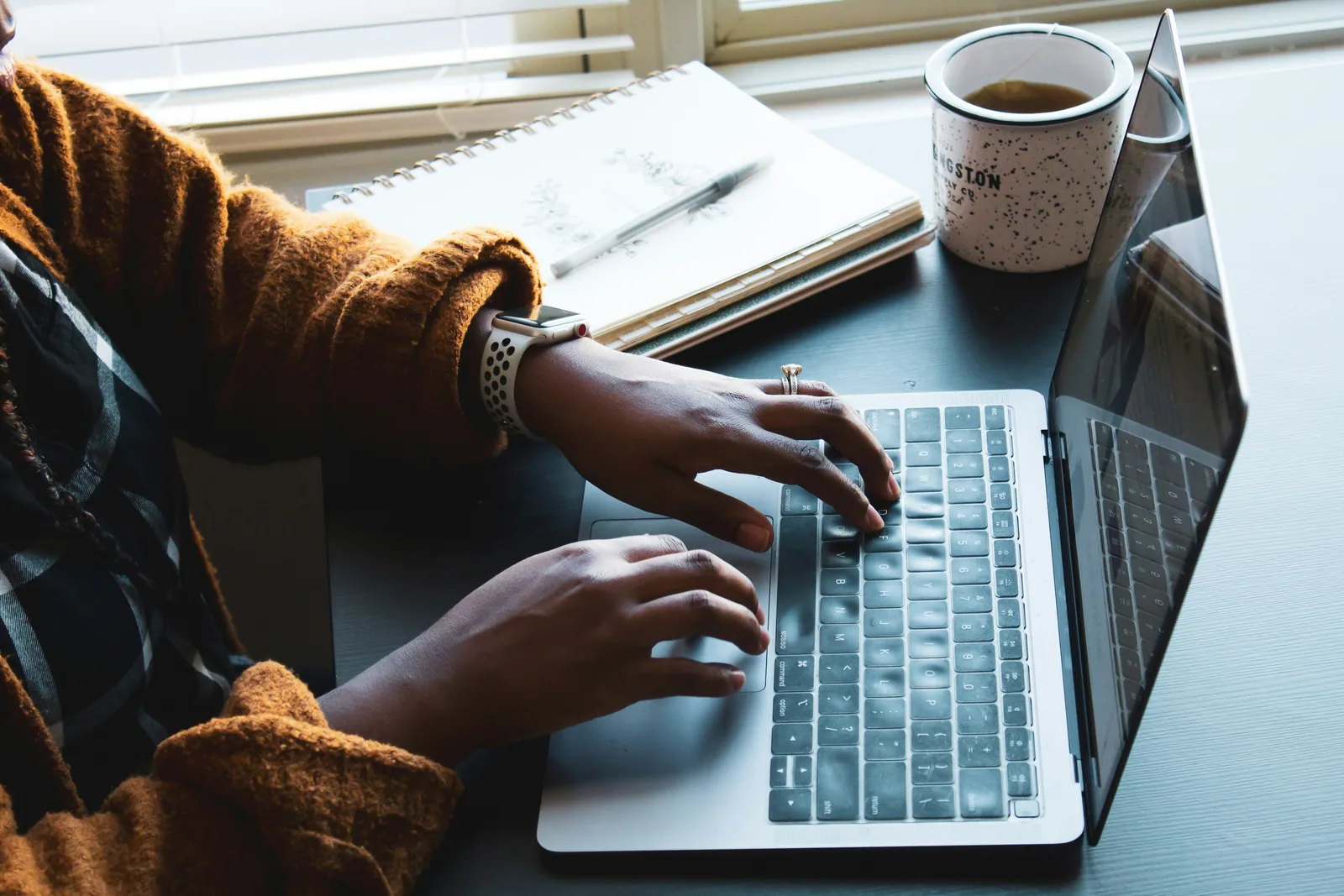 Close-up of hands typing on a laptop by a window, a mug beside it