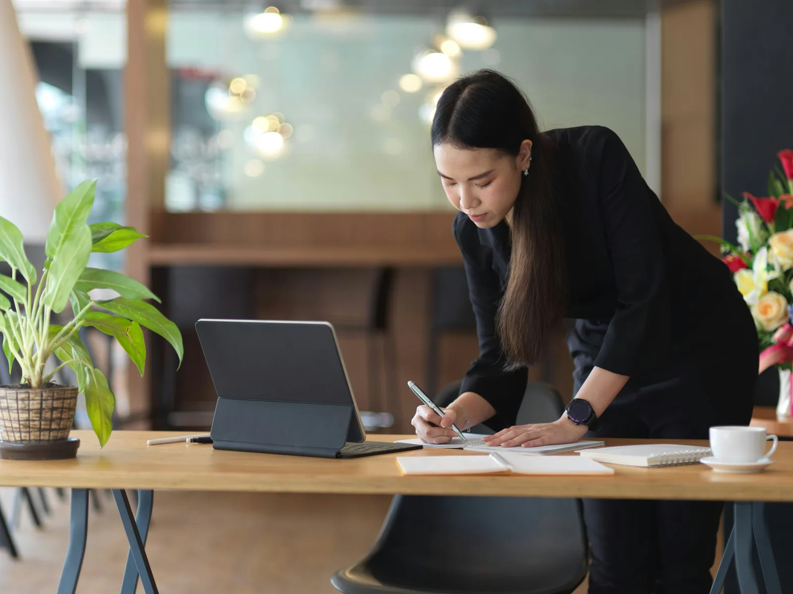 A woman writing at a standing desk beside a laptop, plants behind
