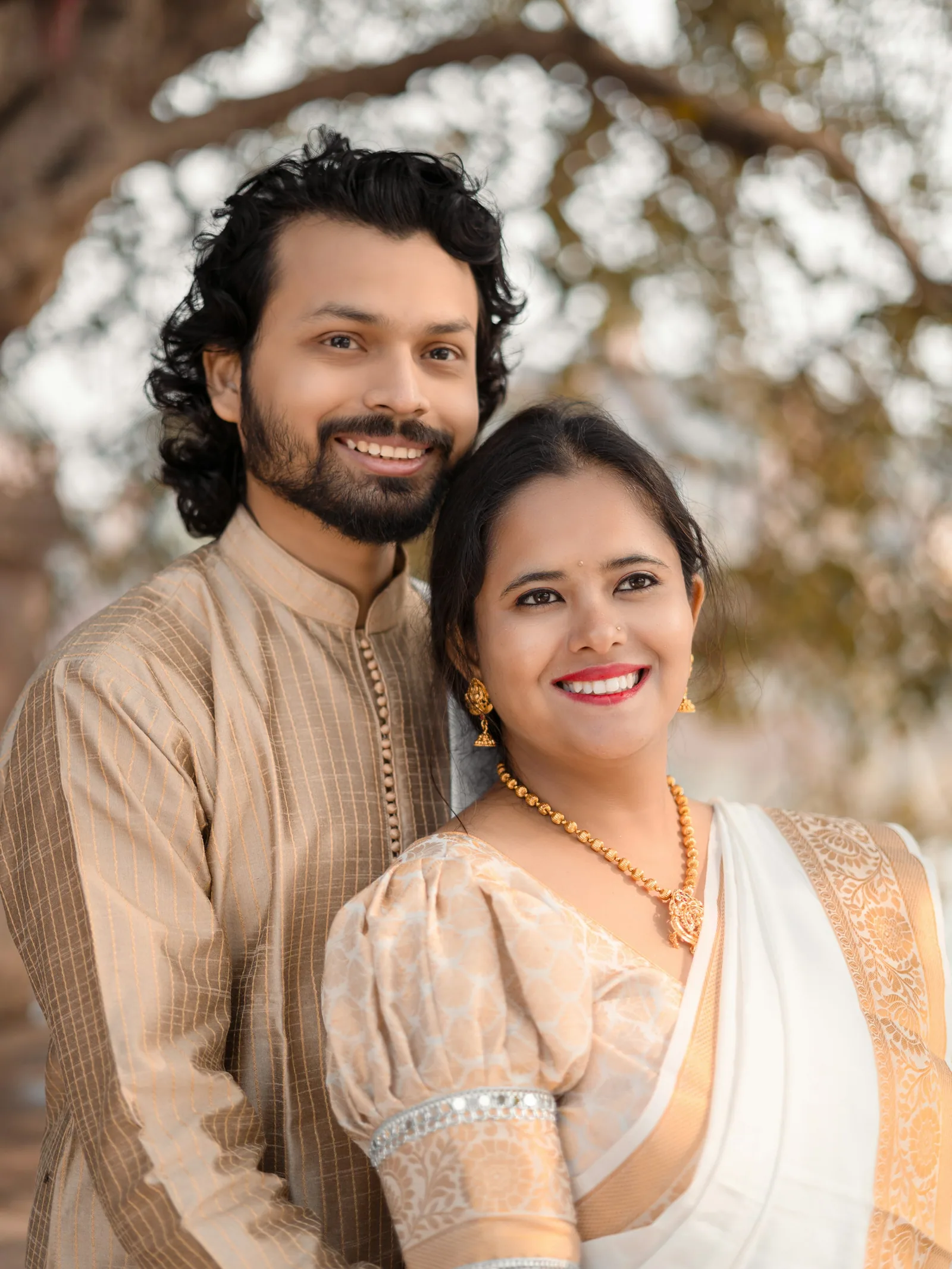 A smiling couple in cream and gold traditional clothes, outdoors