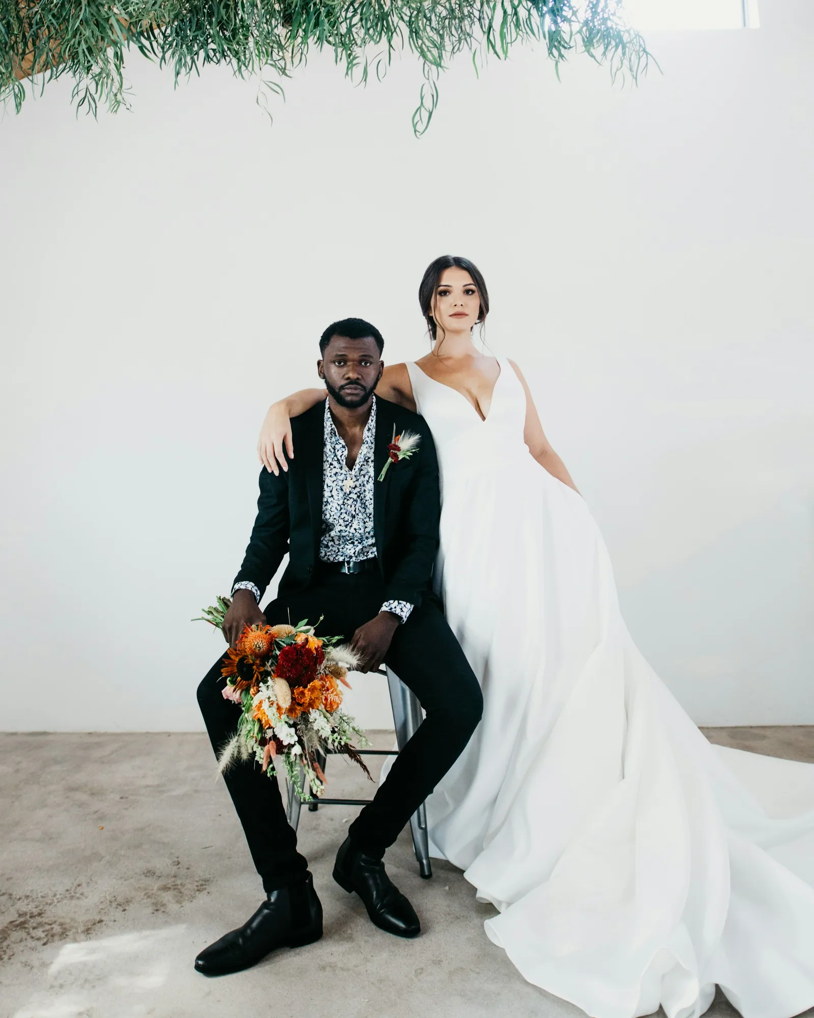 A couple in a white studio, the groom seated with a bouquet, the bride beside him