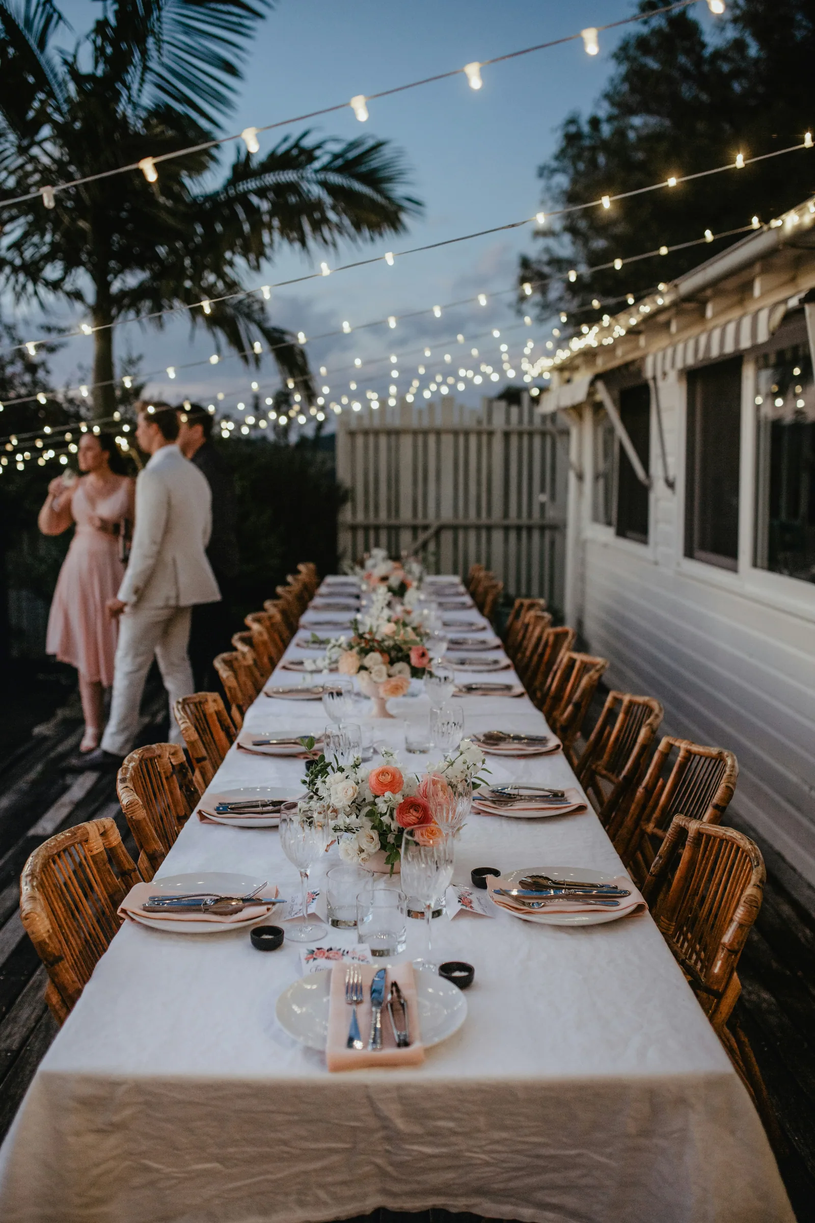 A long outdoor dinner table under string lights, the couple standing at the far end